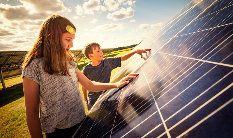 Zwei Kinder stehen vor einem Solarzellenpanel und berühren es. 