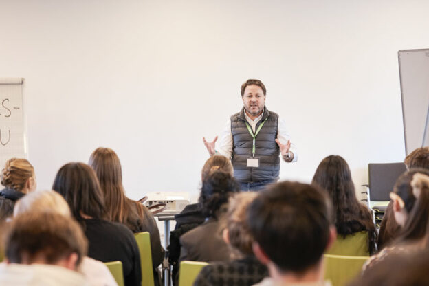 Daniel Harrich steht vor einer sitzenden Gruppe junger Menschen und hält einen Vortrag.