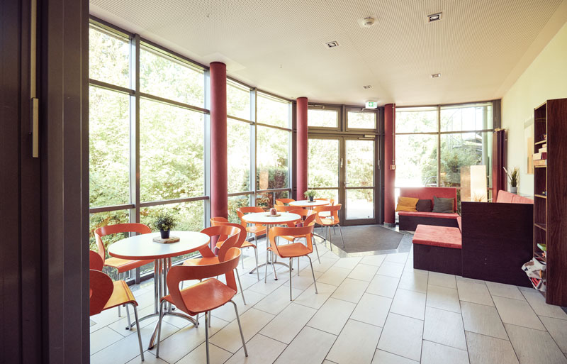 Heller Aufenthaltsraum mit großen Fenstern, mehreren runden Tischen und orangefarbenen Stühlen, mit Blick ins Grüne.