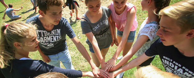 Eine Gruppe von acht Jugendlichen steht im Kreis auf einer Wiese. Sie strecken alle ihre rechte Hand in die Mitte, sodass sich ihre Hände in einem symbolischen Teamkreis berühren. Alle blicken motiviert oder lächelnd auf die Hände. Im Hintergrund sind weitere Jugendliche auf dem Gras zu sehen. Die Szene spielt bei sonnigem Wetter im Freien und vermittelt Gemeinschaftsgefühl und Teamgeist.