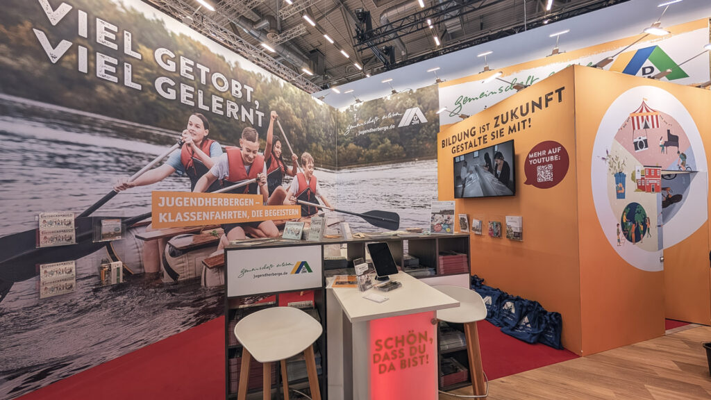 Messestand der Jugendherbergen mit großer Wandgrafik von paddelnden Jugendlichen, Infotheke mit Broschüren und orangefarbener Präsentationsfläche mit Bildschirm.