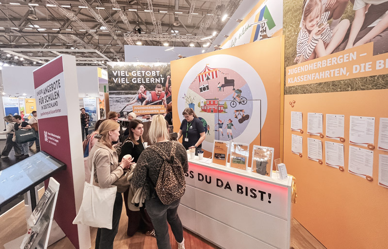 Messestand der Jugendherbergen auf einer Messe, mehrere Besucherinnen im Gespräch am Tresen mit Informationsmaterial und großen orangefarbenen Werbewänden im Hintergrund.