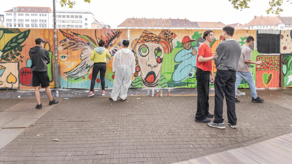 Jugendliche gestalten gemeinsam ein großes Wandbild mit bunten Graffiti-Motiven auf einer Holzfläche im Freien. Einige sprühen aktiv, andere tauschen sich aus oder betrachten das entstehende Kunstwerk. Die Wand zeigt farbenfrohe Figuren, Muster und Symbole.