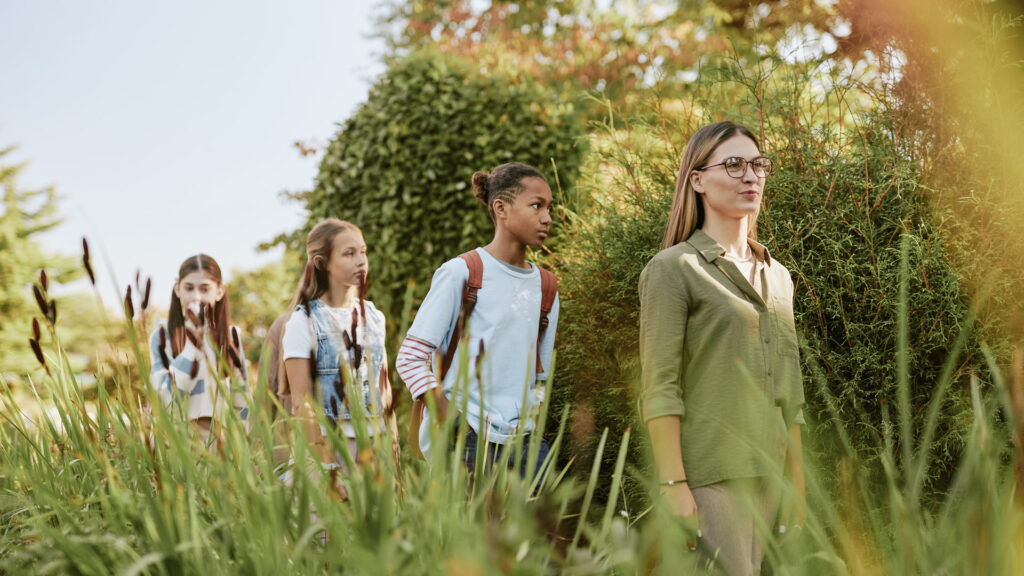 BildungPLUS: Lehrerin geht mit einer Gruppe von Schüler*innen durch eine grüne, naturnahe Umgebung.