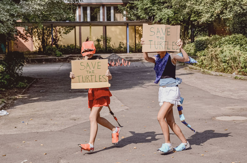 Zwei Kinder laufen über einen Weg. Jedes hält ein Plakat in der Hand auf dem "Save the Turtles" steht. 