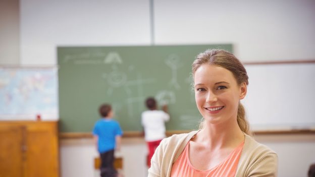Eine Lehrerin vor einer Tafel an der 2 Schüler*innen stehen.