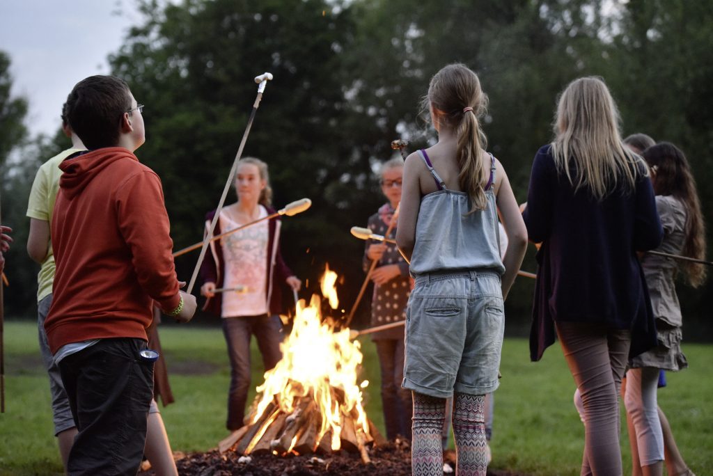 Cirka 8 Kinder stehen um ein Lagerfeuer auf einer Wiese. Sie haben Stäbe mit Stockbrot in der Hand die sie über das Lagerfeuer halten.