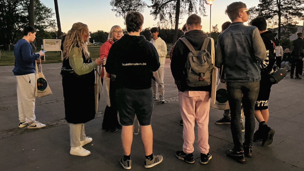 Eine Gruppe Jugendlicher und Erwachsener steht bei Dämmerung auf einem Platz und bereitet sich auf eine geführte Erlebnistour vor. Einige tragen Taschen, im Hintergrund sind Bäume und weitere Teilnehmende zu sehen.