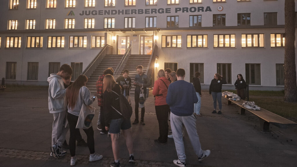 Eine Gruppe Jugendlicher und Erwachsener steht bei Dämmerung vor der hell erleuchteten Jugendherberge Prora und bereitet sich auf eine gemeinsame Erlebnistour vor.