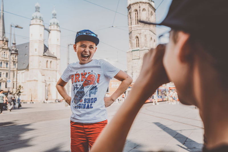 Ein lachender Junge mit Basecap und Sonnenbrille wird fotografiert und steht vor einer Kirche.