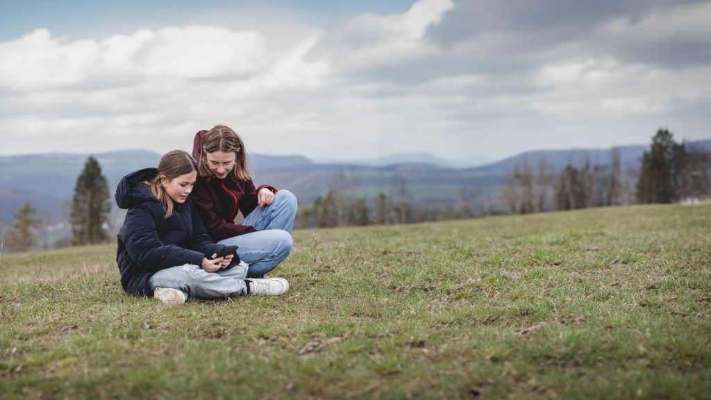 Natur-Erlebispädagogik Programm in Lauterbach. Zwei Mädchen sitzen auf einer Wiese und schauen auf ein Handy.
