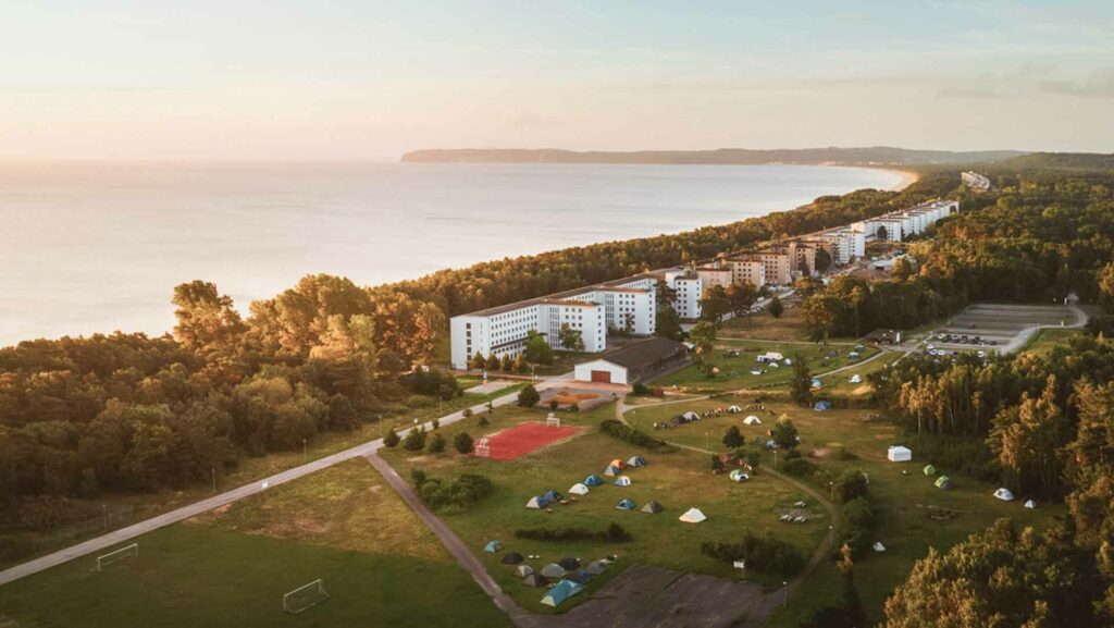 Luftaufnahme eines Zeltplatzes in Prora bei Sonnenuntergang. Zwischen Ostsee, Wald und den langen Gebäuden stehen mehrere Zelte auf einer großen Wiese.