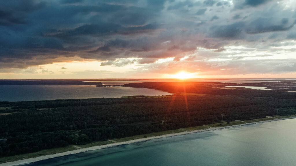 Luftaufnahme der Küste von Prora bei Sonnenaufgang. Die Sonne steht tief am Horizont über Wald, Wasserflächen und Strand, während dunkle Wolken den Himmel prägen.