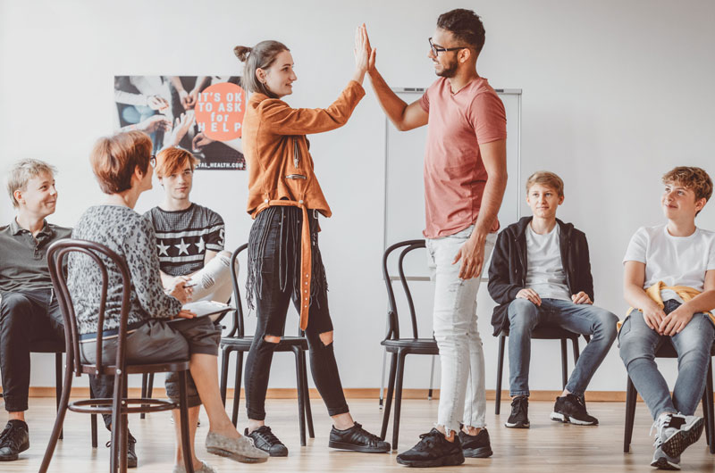 Ein Gruppe von Schülern:Innen sitzt in einem Raum mit Stühlen. 5 Schüler:Innen sitzen. Zwei Schüler:Innen stehen und klatschen ab.